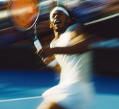 Tennis player in white attire swinging racket on a blue tennis court in motion blur.